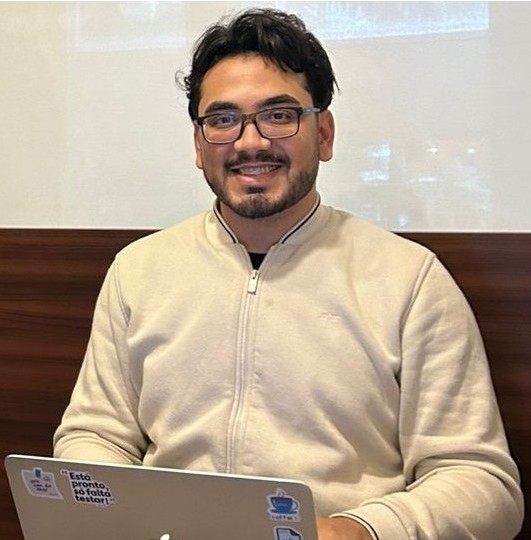 Foto de perfil de Vinícius Augusto, moreno, óculos, cabelos pretos ondulados, vestindo uma camiseta verde e um casaco preto. Ao fundo um ambiente desfocado.