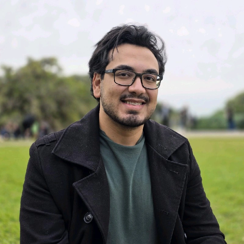 Foto de perfil de Vinícius Augusto, moreno, óculos, cabelos pretos ondulados, vestindo uma camiseta verde e um casaco preto. Ao fundo um ambiente desfocado.