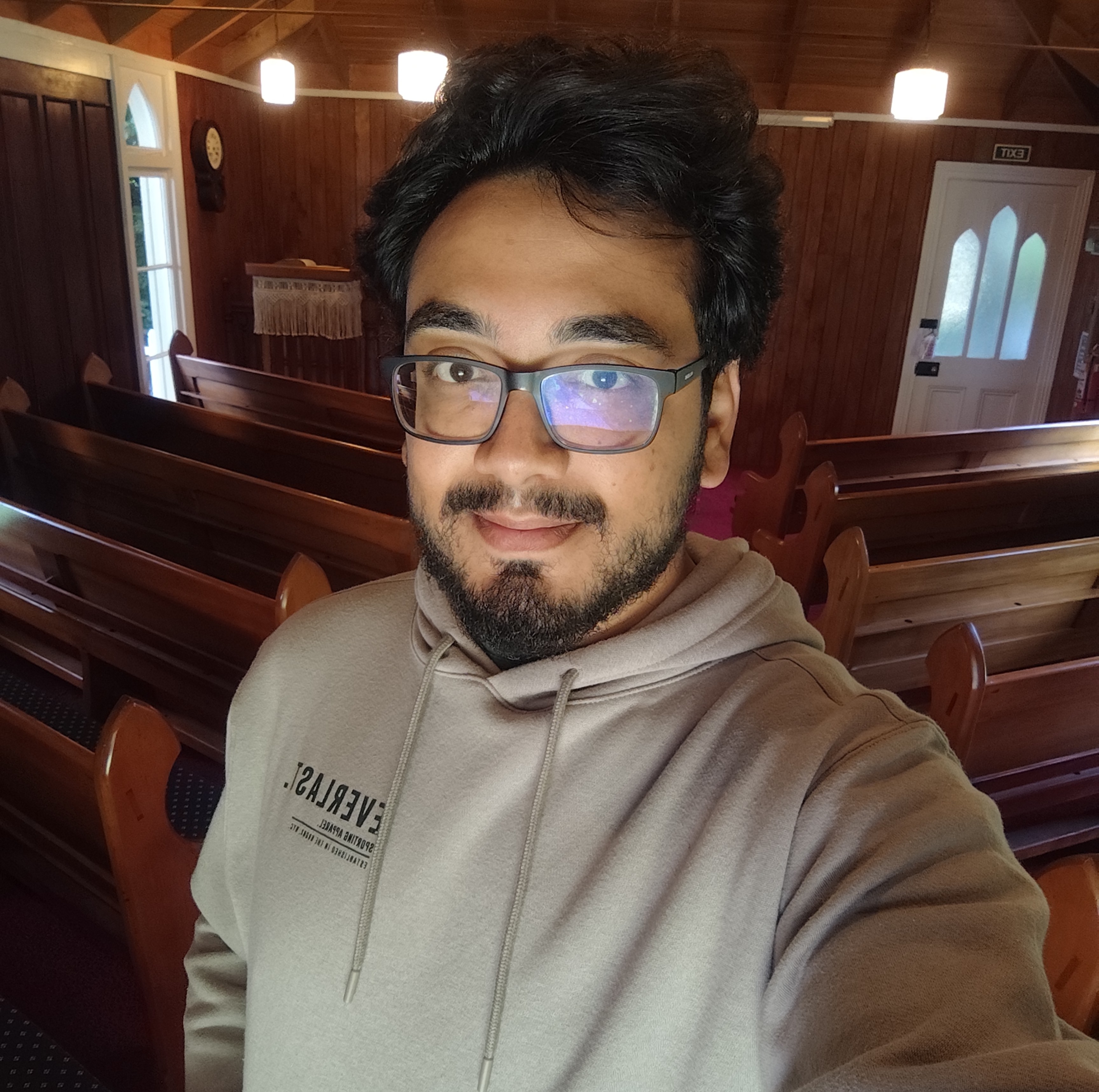 Foto de perfil de Vinícius Augusto, moreno, óculos, cabelos pretos ondulados, vestindo uma camiseta verde e um casaco preto. Ao fundo um ambiente desfocado.